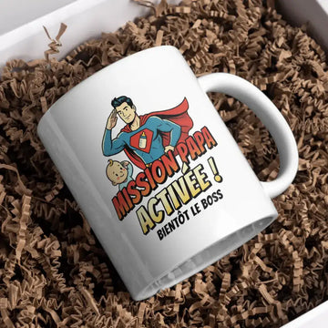 Tasse en céramique blanche ornée d'une illustration cartoon colorée de Superman saluant tout en tenant un biberon, avec le texte « mission papa activée ! Bientôt le boss » imprimé en lettres grasses orange et jaunes.