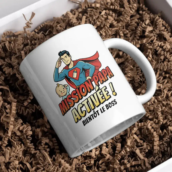 Tasse en céramique blanche ornée d'une illustration cartoon colorée de Superman saluant tout en tenant un biberon, avec le texte « mission papa activée ! Bientôt le boss » imprimé en lettres grasses orange et jaunes.